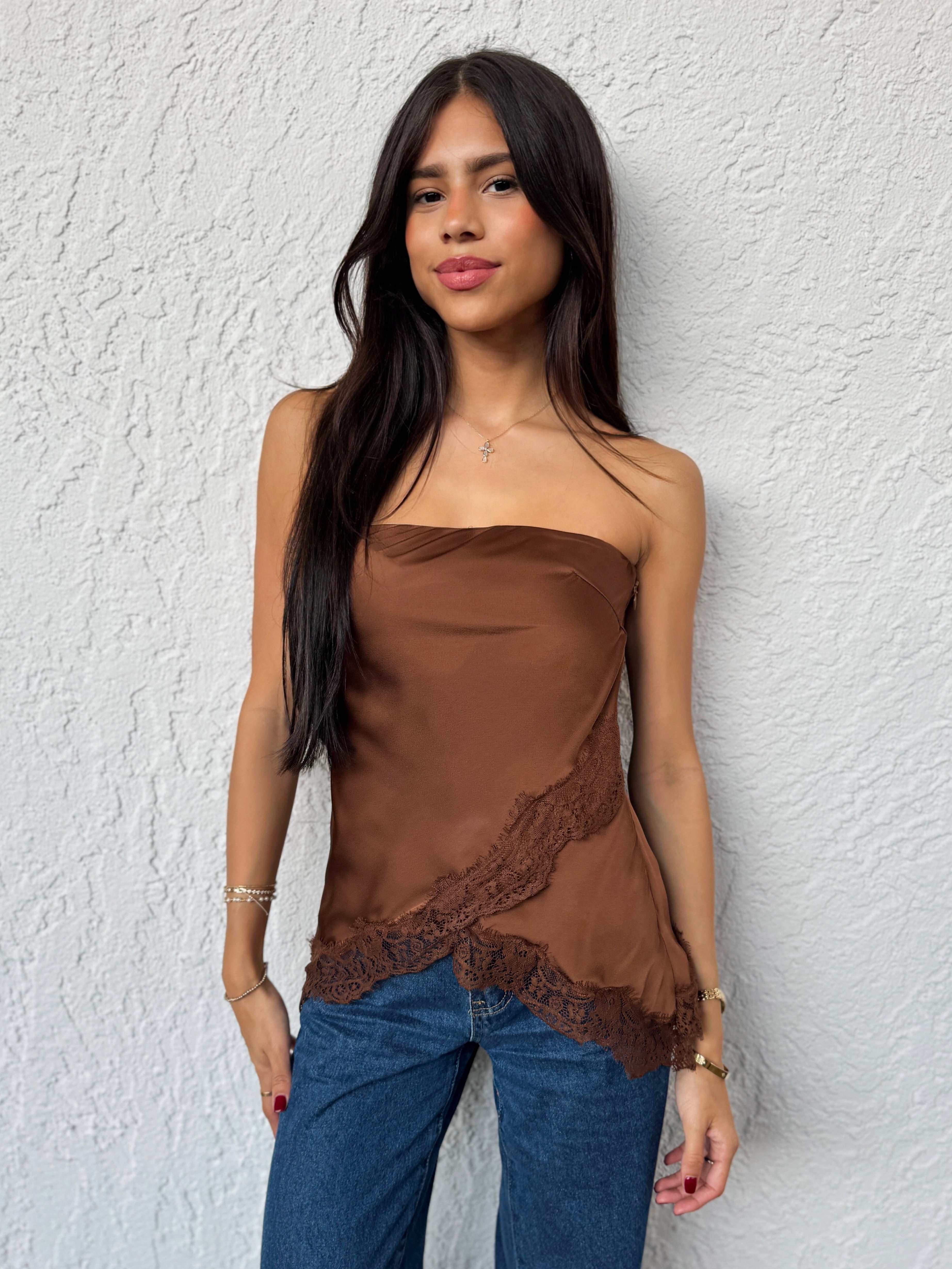 Woman wearing a brown strapless top with lace details and blue jeans against a white wall.